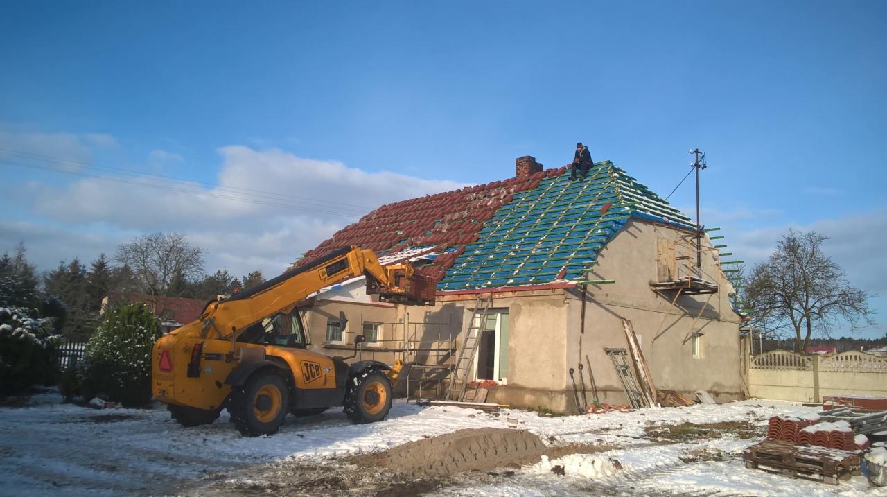 Częściowo zdjęty dach z widoczną konstrukcją i nowymi dachówkami, z żółtą maszyną budowlaną JCB z przodu i pracownikiem na dachu, zimowy krajobraz.
