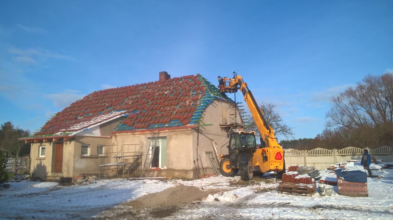 Dom z częściowo zdjętym, czerwonym dachem, robotnicy na podnośniku układają nowe dachówki, w tle błękitne niebo i śnieg na ziemi.