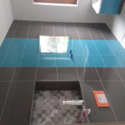 Wykończona ściana w łazience z szarymi i turkusowymi płytkami, widoczne narzędzia glazurnicze, poziomica i resztki materiałów budowlanych w otworze na umywalkę.