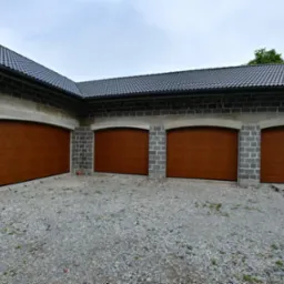 Cztery brązowe segmentowe bramy garażowe w szarym, surowym budynku z bloczków betonowych pod ciemnym dachem z dachówki, widok z zewnątrz na żwirowy podjazd.