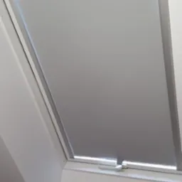 Okno dachowe z szarą roletą w minimalistycznym, białym wnętrzu, widoczne z bliskiej perspektywy.