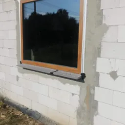 Montaż okna z drewnianą ramą w bloku z betonu kom&oacute;rkowego w Raciborzu, widoczny parapet zewnętrzny i ślady po zaprawie.