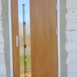 Brązowe drzwi zewnętrzne z pionowym przeszkleniem, zamontowane w surowym otworze ściennym z białego betonu komórkowego, zabezpieczone folią ochronną na ościeżnicy.