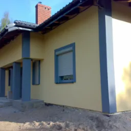 Żółty dom jednorodzinny z szarą stolarką okienną, kolumnami i ceglanym kominem, widoczny z boku, teren wokół domu jeszcze niezagospodarowany.