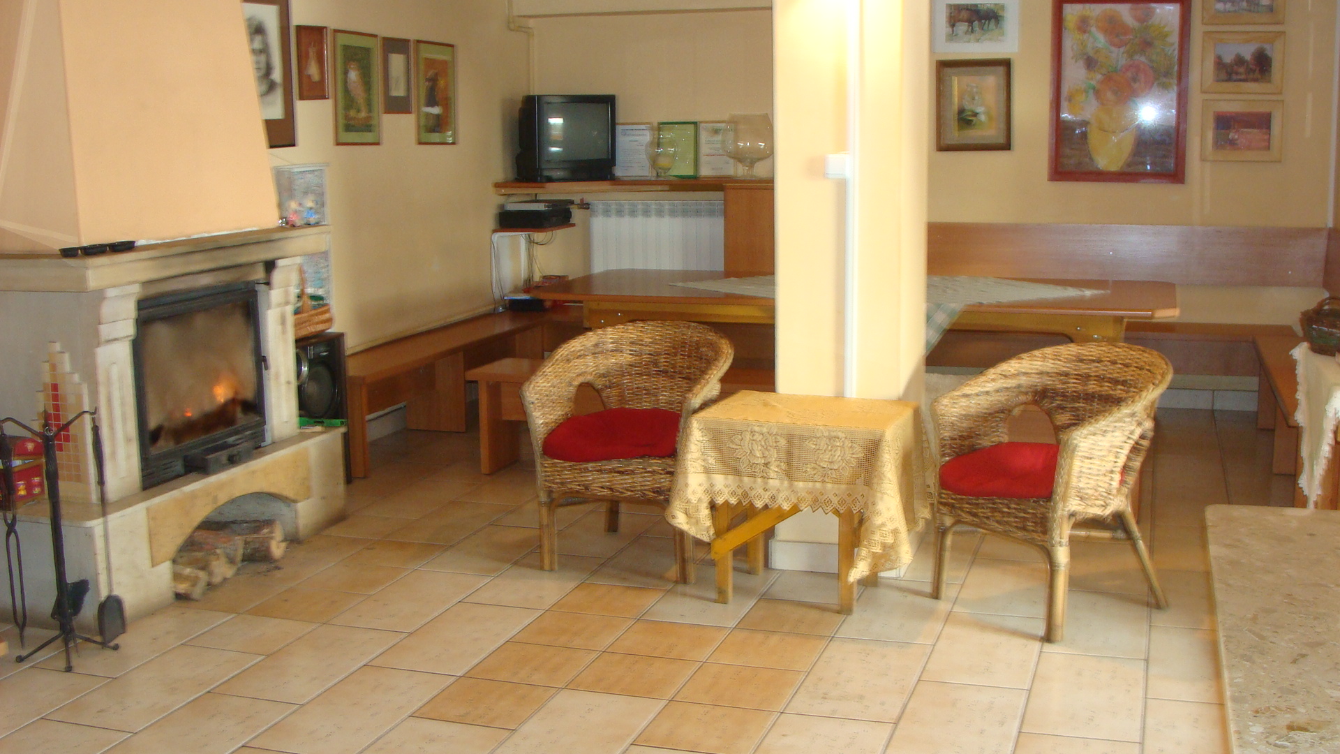 Przytulne wnętrze pokoju z kominkiem, wiklinowymi fotelami z czerwonymi poduszkami i drewnianymi ławami, udekorowane obrazami na ścianach.