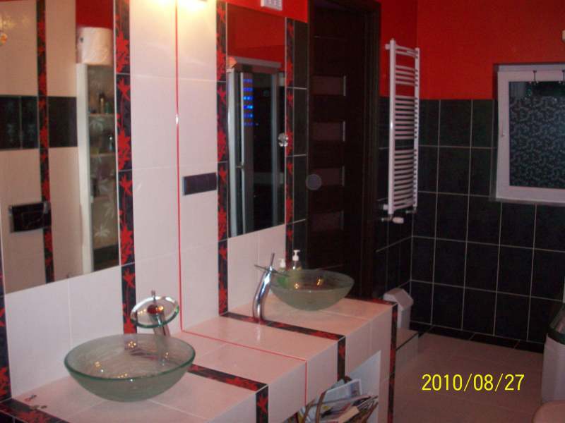 Nowoczesna łazienka z czerwonymi akcentami: szklane umywalki na blacie z białych płytek z dekoracyjną bordiurą, czarne płytki na ścianie z grzejnikiem drabinkowym, ciemne drzwi.