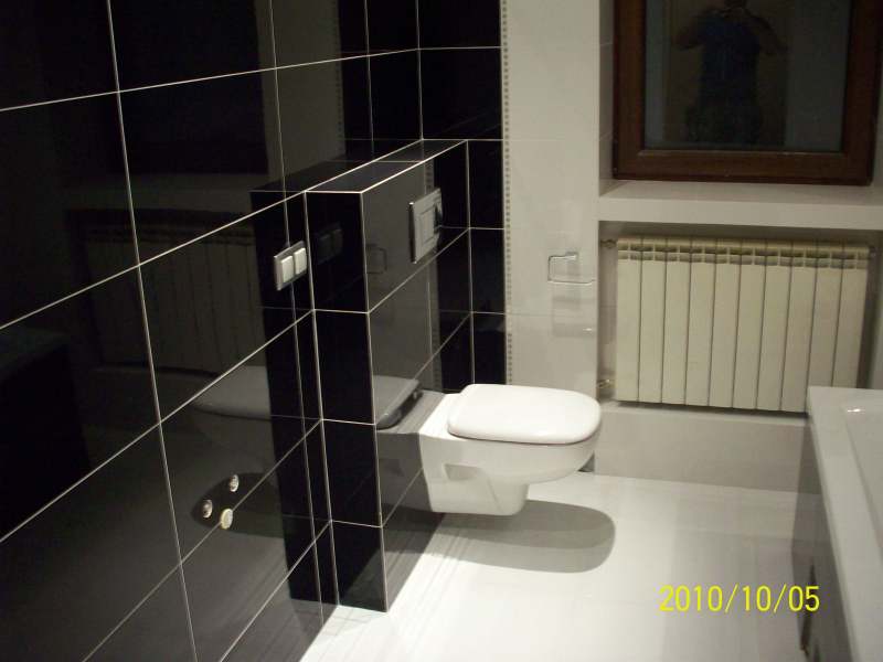 Nowoczesna łazienka z czarnymi, błyszczącymi płytkami i podwieszaną toaletą; widoczny grzejnik i odbicie w płytkach.