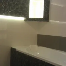 Nowoczesna łazienka z wanną obłożoną mozaiką, podświetlane lustro z mozaikową ramą, białe płytki ścienne z szarym pasem.