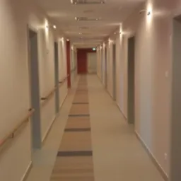 Długi korytarz w budynku biurowym z beżowymi ścianami, szarymi drzwiami i drewnianą poręczą, oświetlony lampami ściennymi i sufitowymi, z wykładziną podłogową w pasy.