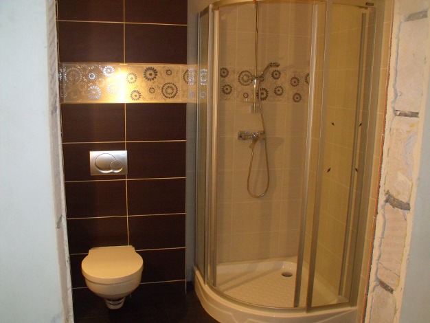 Wykończona łazienka z brązowymi płytkami, dekoracyjnym pasem z motywami kwiatowymi, podwieszaną toaletą i kabiną prysznicową z półokrągłymi drzwiami przesuwnymi.