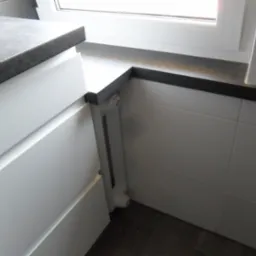 Szary blat z widoczną fakturą kamienia, zamontowany w rogu przy oknie, połączony z białymi szafkami kuchennymi i grzejnikiem.