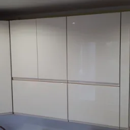 Jasne, lakierowane szafki kuchenne na wymiar, z subtelnymi, poziomymi uchwytami, zamontowane w rogu kuchni z żółtą ścianą w tle.