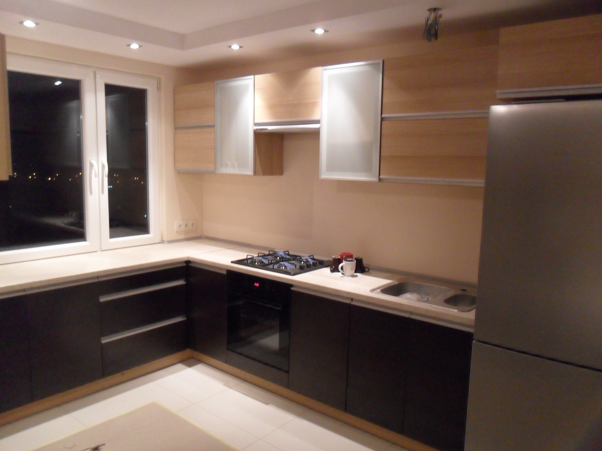 Nowoczesna kuchnia z ciemnymi szafkami dolnymi i jasnymi górnymi, widok na płytę grzewczą, piekarnik, zlew i lodówkę ze stali nierdzewnej.