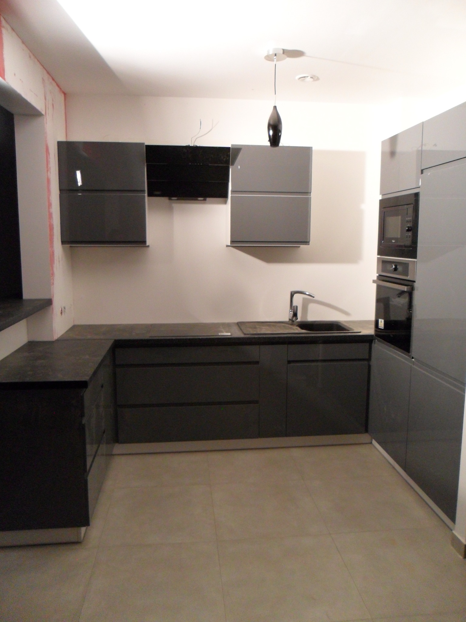 Nowoczesna, szara zabudowa kuchenna z ciemnym blatem i sprzętem AGD w trakcie montażu, widoczne niedokończone prace wykończeniowe na ścianie.