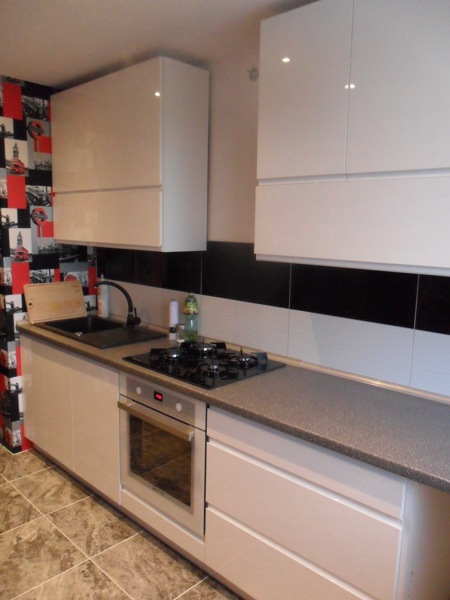 Nowoczesna kuchnia z białymi szafkami wiszącymi, szarym blatem, płytą gazową i piekarnikiem; na ścianie tapeta w motywy londyńskie.