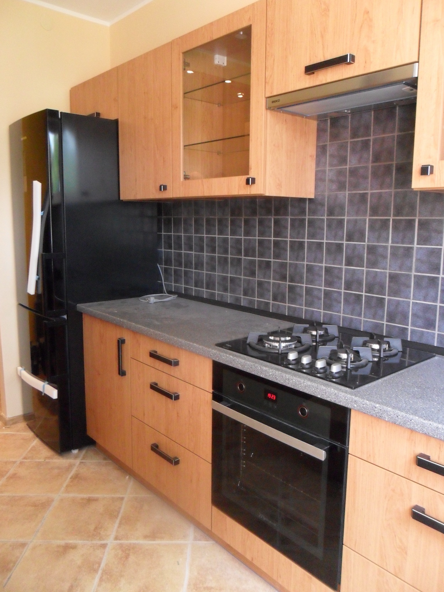 Jasna kuchnia z drewnianymi frontami, czarnym sprzętem AGD i grafitowym blatem, widoczny piekarnik z wyświetlaczem, płyta gazowa i czarna lodówka.