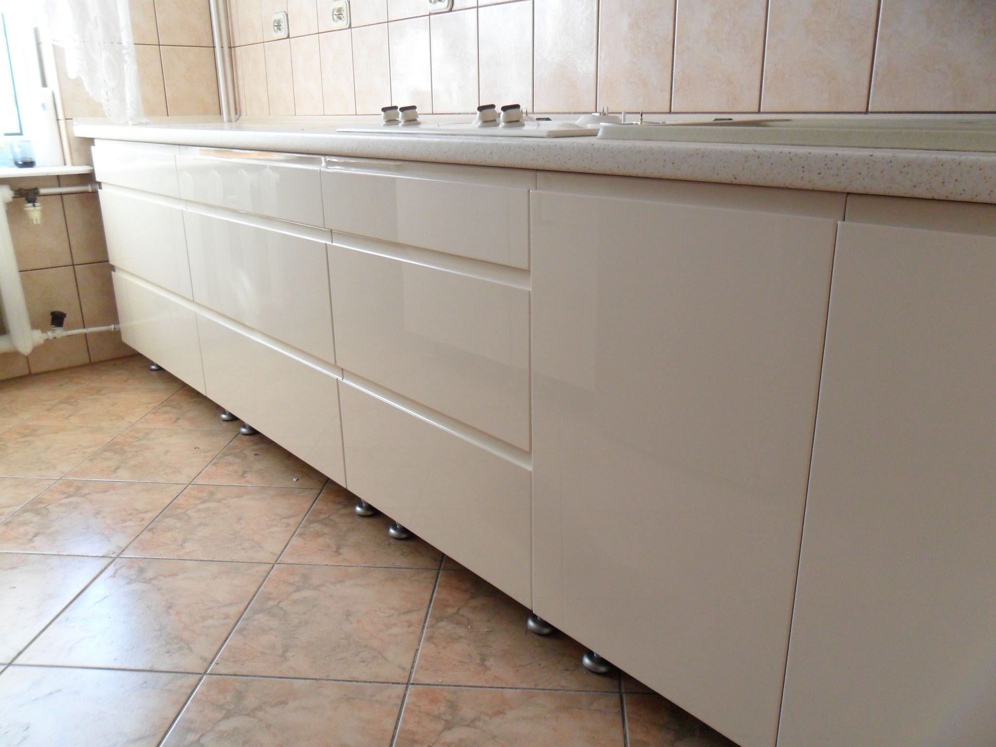 Beżowe, lakierowane szafki kuchenne z szufladami i blatem w tonacji beżu, widoczne od dołu, z ceramiczną płytą grzewczą na blacie i beżowymi płytkami na ścianie oraz podłodze.