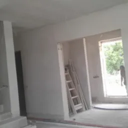 Wnętrze w stanie surowym po tynkowaniu maszynowym: widoczne gładkie, szare ściany, sufit, schody z pustaków, drewniana drabina oparta o ścianę przy przejściu do kolejnego pomieszczenia, wystające...