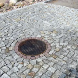 Szara kostka granitowa ułożona wokół studzienki kanalizacyjnej z czerwoną obwódką, widok z góry.