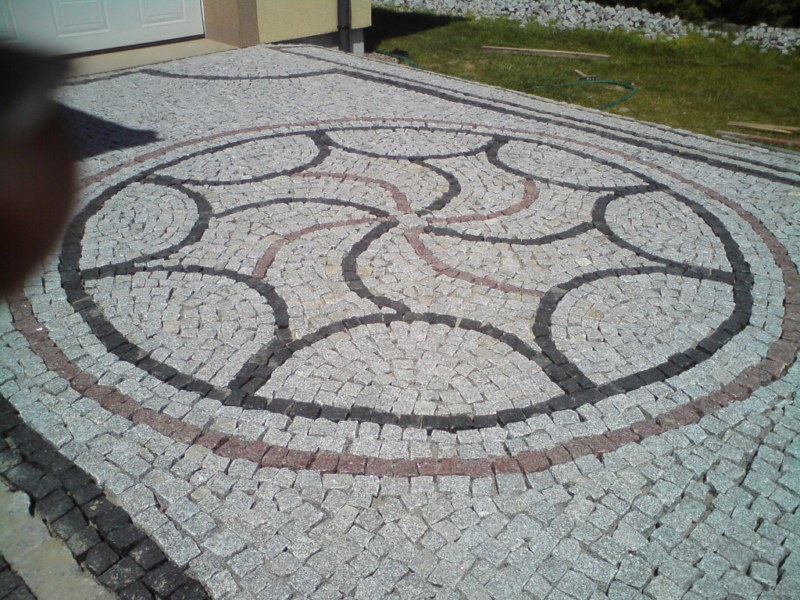 Ułożona kostka granitowa tworząca okrągły wzór na podjeździe, z elementami w kolorze szarym, czarnym i ceglastym.