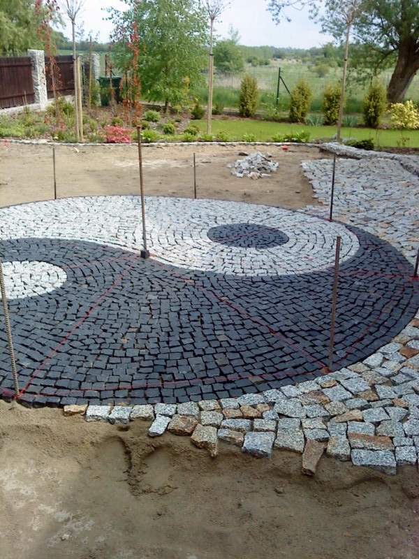 Kompozycja Yin Yang ułożona z czarnej i szarej kostki granitowej na etapie realizacji, widoczne sznurki wyznaczające kształt, w tle ogród i drzewa.