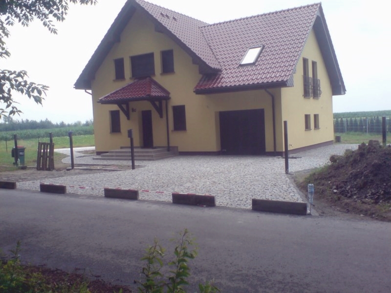 Ułożona nawierzchnia z szarej kostki granitowej przed domem z żółtą elewacją i czerwoną dachówką, widoczne obrzeża i fragment drogi asfaltowej.