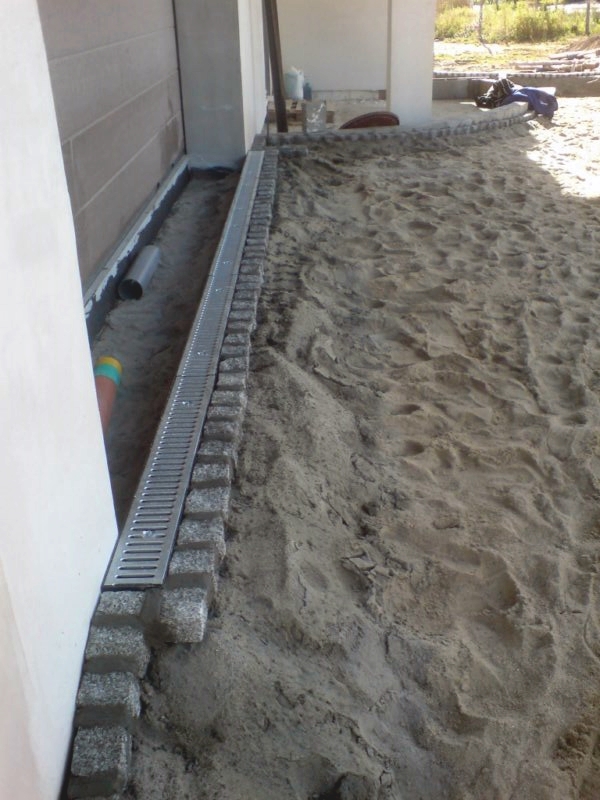 Montaż liniowego odwodnienia posesji z kostki granitowej na podsypce piaskowej, widoczne rury odprowadzające wodę.