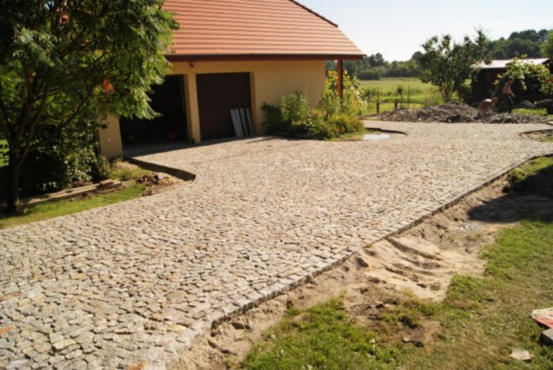 Ułożona nawierzchnia z szarego łupka granitowego prowadząca do garażu, widoczne obrzeża i przygotowane podłoże po bokach.