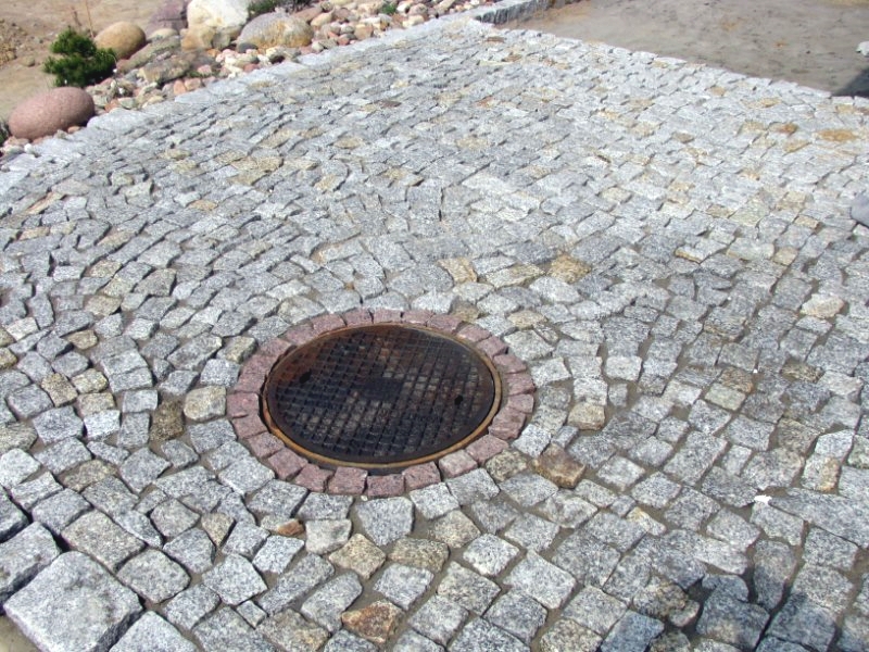 Szara kostka granitowa ułożona wokół studzienki kanalizacyjnej z czerwoną obwódką, widok z góry.