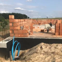 Wznoszone ściany parteru z czerwonej cegły na fundamencie, widoczne niebieskie rury instalacji w wykopie, w tle krajobraz z lasem i polem pod błękitnym niebem.