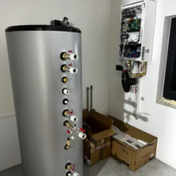 Szary, cylindryczny zbiornik buforowy z licznymi przyłączami na ścianie w pomieszczeniu, obok otwarta obudowa sterownika z widocznymi podzespołami elektronicznymi i przewodami, oraz kartony po...