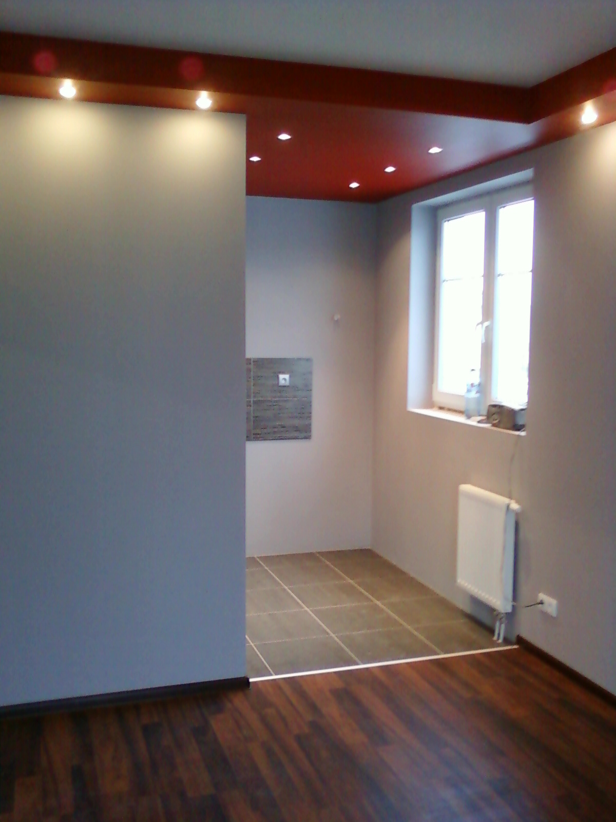 Wykończone wnętrze: szare ściany, drewniana podłoga przechodząca w kafelki w łazience, czerwony sufit z oświetleniem punktowym oraz biały grzejnik pod oknem.