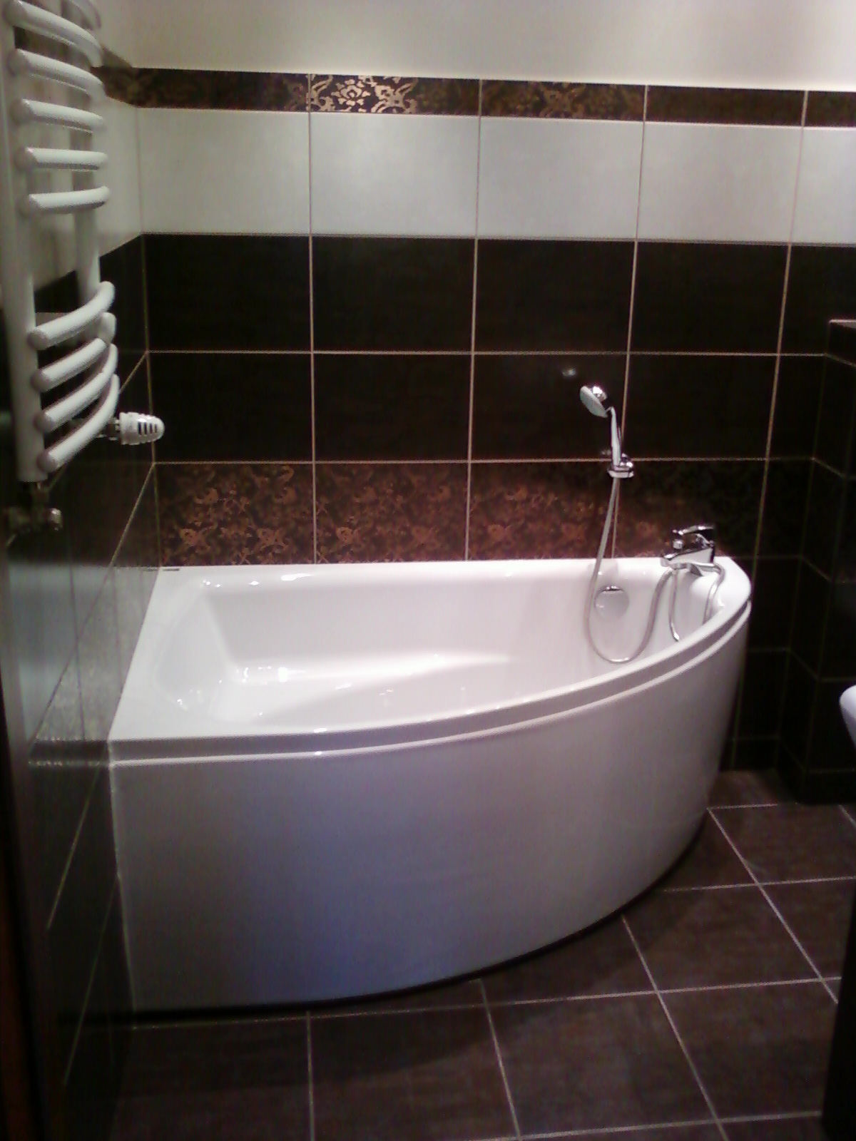 Nowoczesna, narożna wanna w wykończonej łazience z brązowymi i białymi płytkami oraz grzejnikiem drabinkowym.