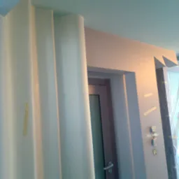 Gładź bezpyłowa na zaokrąglonej kolumnie i ścianie w trakcie prac wykończeniowych, widoczna taśma malarska i zabezpieczone drzwi.