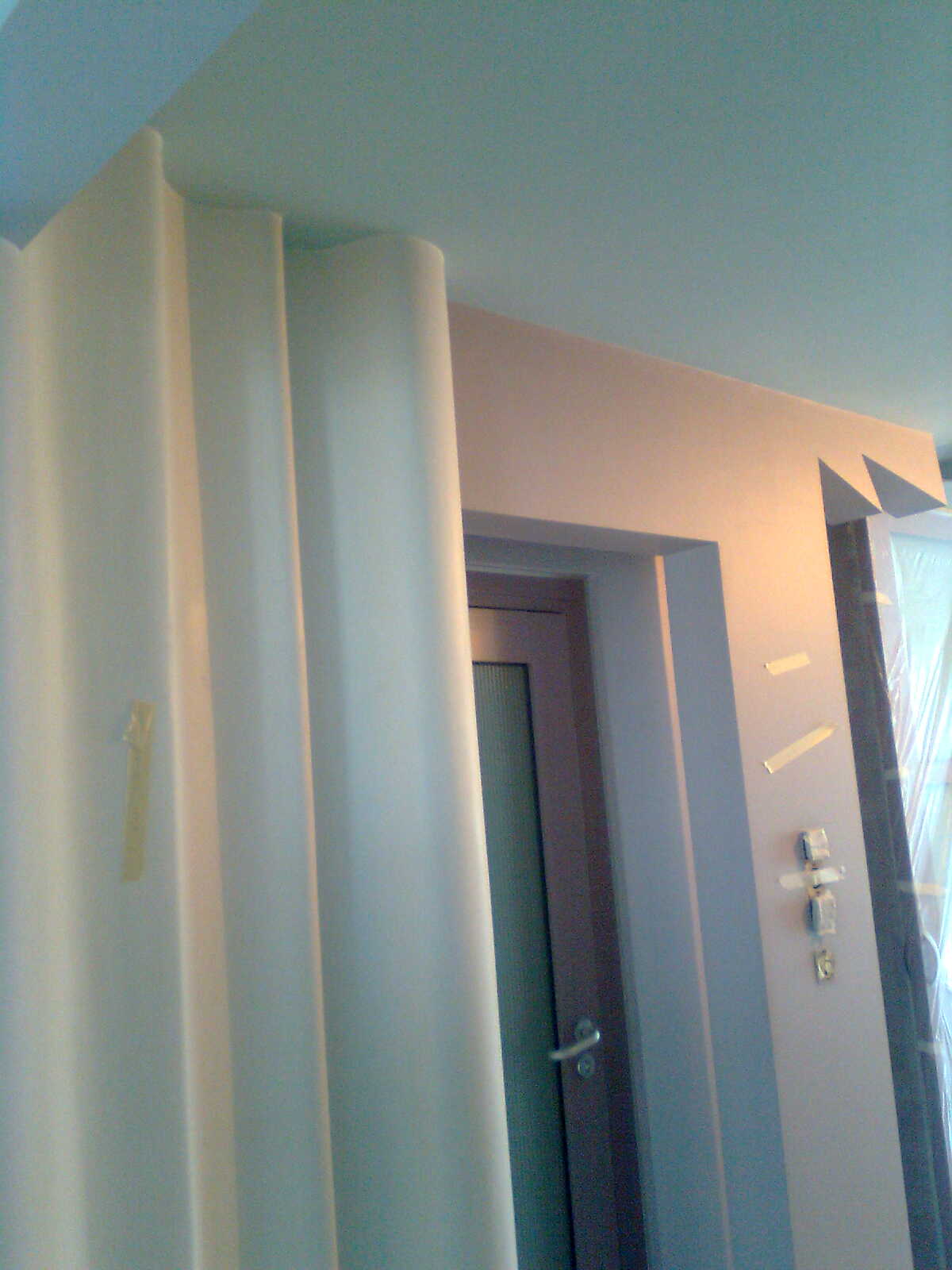 Gładź bezpyłowa na zaokrąglonej kolumnie i ścianie w trakcie prac wykończeniowych, widoczna taśma malarska i zabezpieczone drzwi.