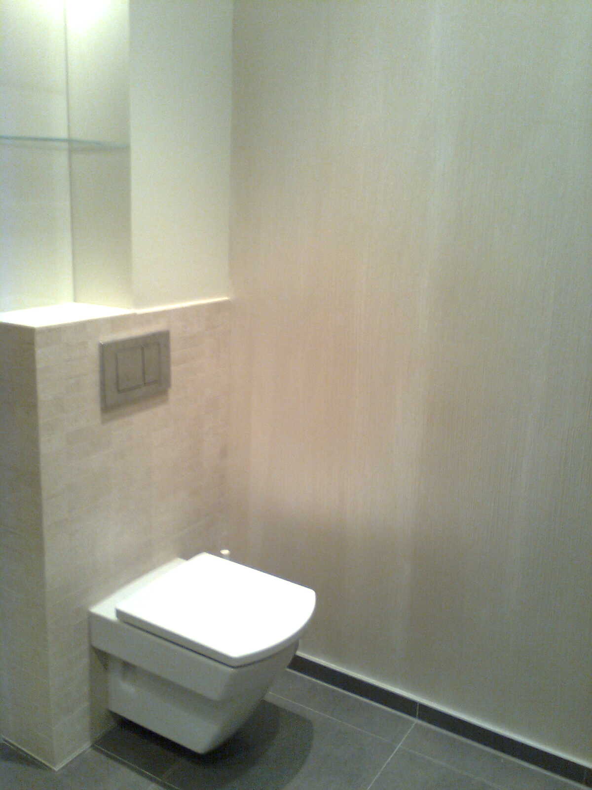 Wisząca toaleta z zabudowaną spłuczką na tle beżowych płytek i szarej podłogi z ciemną listwą przypodłogową, w łazience wykończonej w odcieniach beżu i szarości.