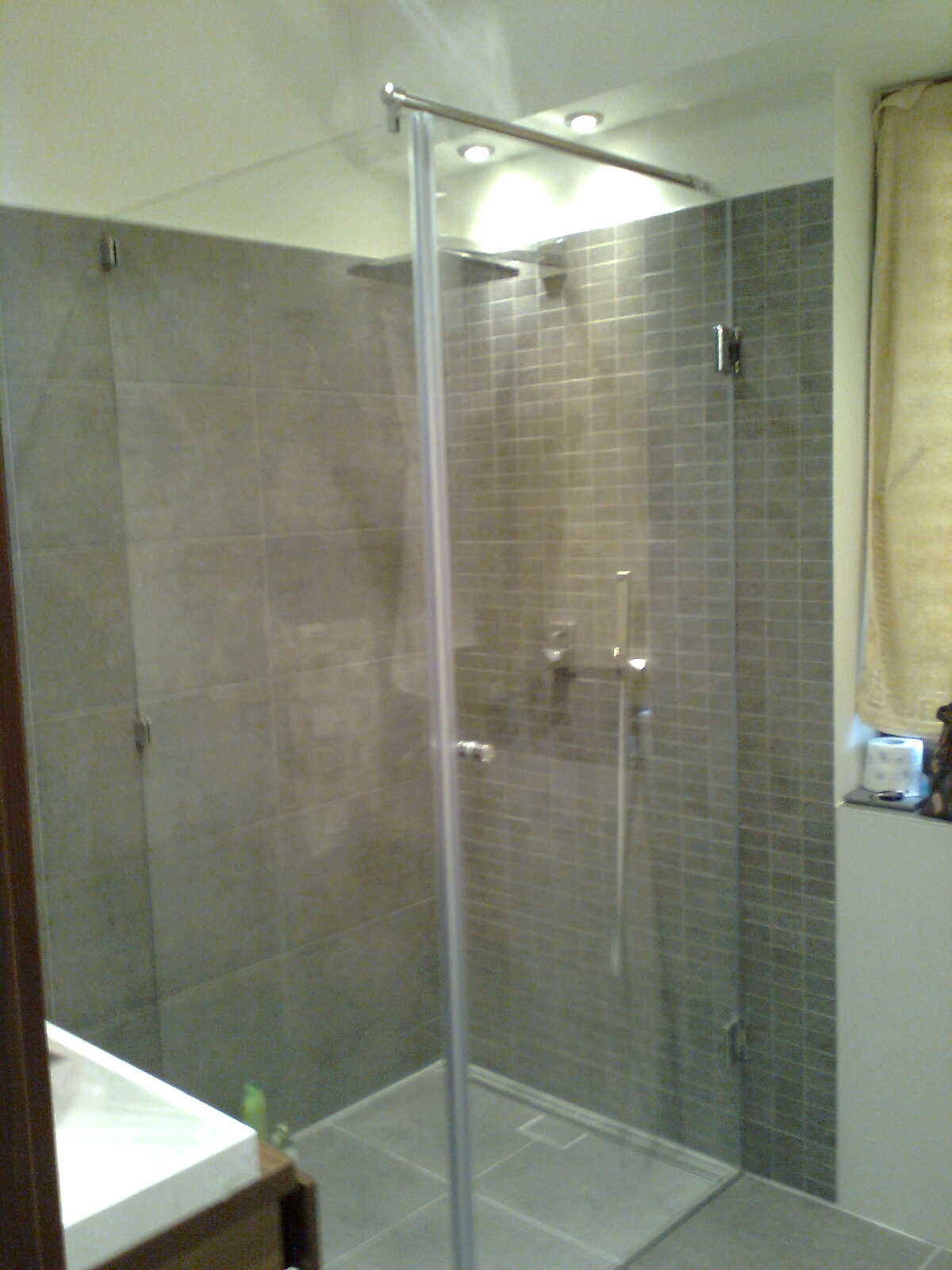 Kabina prysznicowa z szarymi płytkami i szklanymi drzwiami, widoczny chromowany zestaw natryskowy i umywalka z drewnianą szafką w tle.