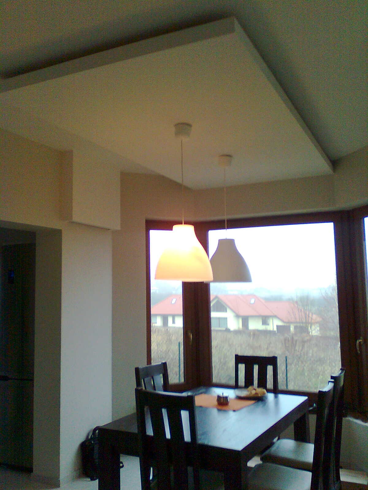 Nowoczesna jadalnia z ciemnym drewnianym stołem i krzesłami, widok na domy z czerwonymi dachami przez okno, dwie lampy wiszące z białymi kloszami, gładź bezpyłowa na suficie.
