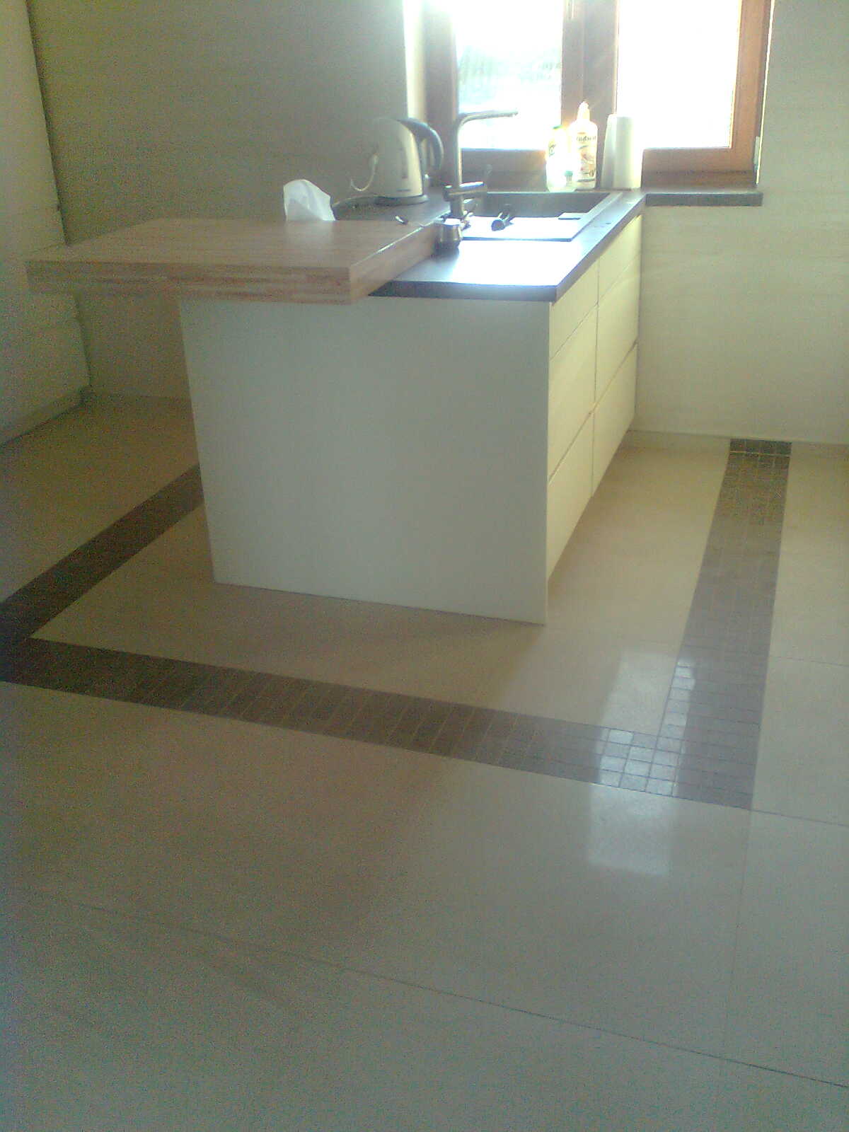 Nowoczesna, minimalistyczna kuchnia z białymi szafkami, ciemnym blatem i drewnianym blatem pomocniczym. Umywalka pod oknem z widocznym czajnikiem i płynem do mycia naczyń. Na podłodze jasne płytki...