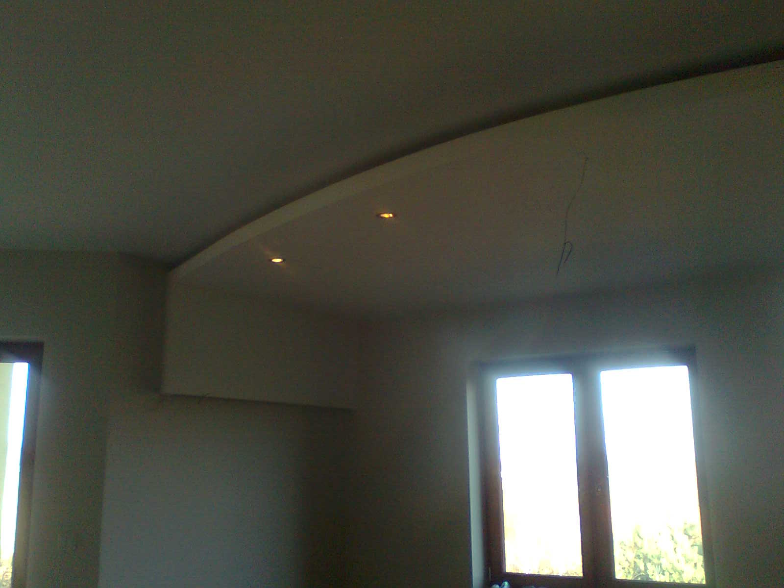 Wykończone wnętrze pokoju z podwieszanym sufitem z oświetleniem punktowym, widok na okno z widoczną zielenią na zewnątrz.