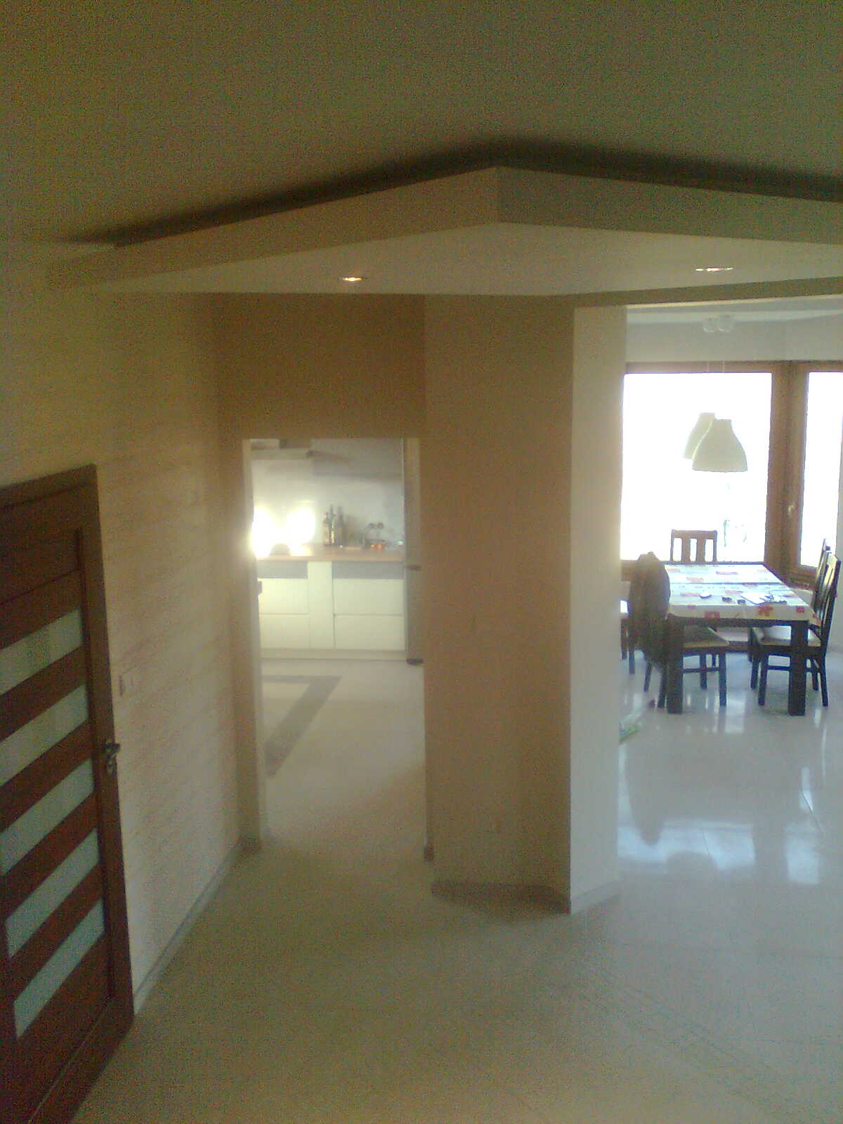 Wnętrze domu po remoncie: widok na kuchnię i jadalnię z drewnianą stolarką okienną, drzwi wewnętrzne z ukośnymi panelami, sufit podwieszany z oświetleniem punktowym.