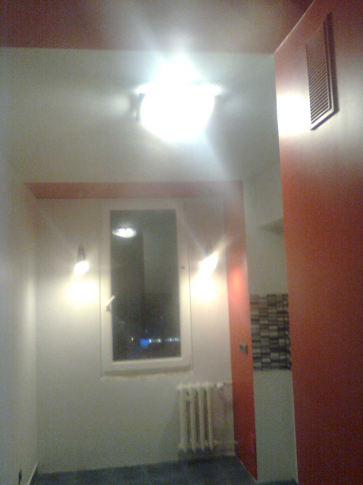 Wnętrze korytarza po remoncie, z widocznym oknem, grzejnikiem i punktowym oświetleniem, ściany pomalowane w odcieniach bieli i czerwieni.