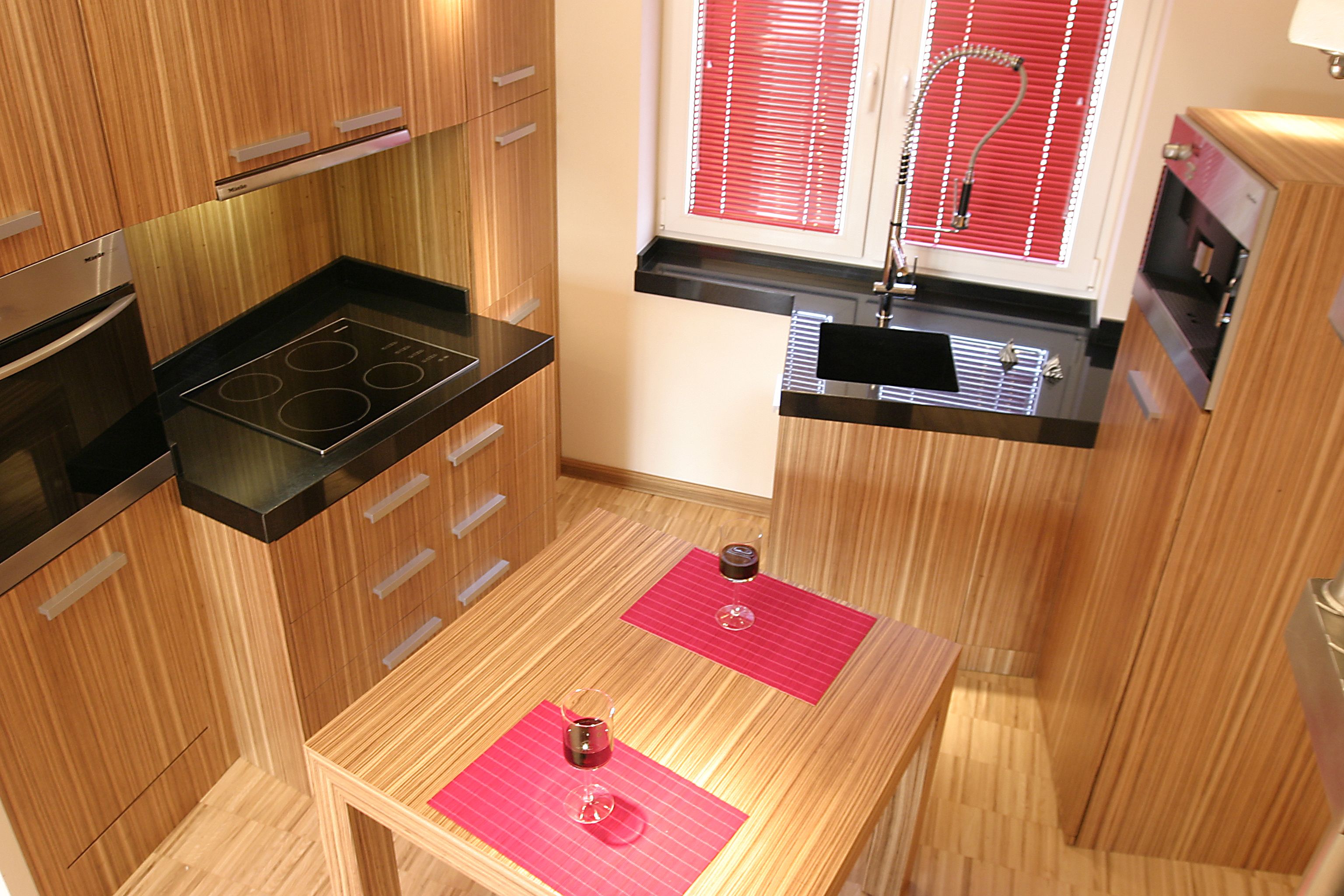 Nowoczesna kuchnia z drewnianymi szafkami, czarnym blatem, płytą indukcyjną i dwoma kieliszkami wina na drewnianym stole z czerwonymi podkładkami.