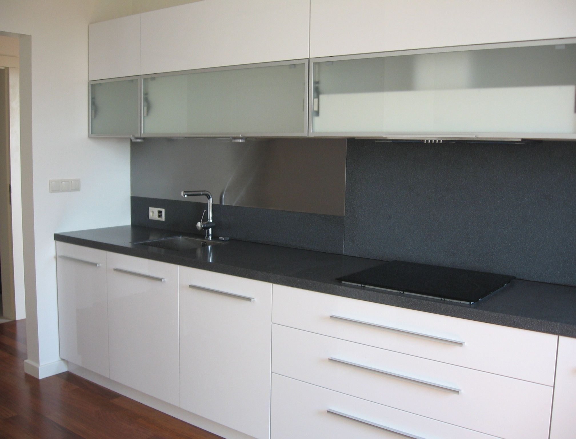 Nowoczesna kuchnia z białymi szafkami na wymiar, czarnym blatem i szklanymi frontami szafek wiszących, z widocznym fragmentem podłogi z ciemnego drewna.