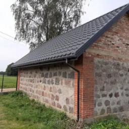 Budynek gospodarczy z nowym, grafitowym dachem z blachodachówki, wykończony czarnymi rynnami, ściany wykonane z kamienia i cegły, widok z zewnątrz na tle drzew i pola.