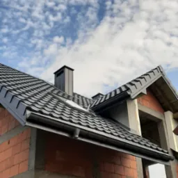 Fragment dachu z ciemnej blachodachówki z kominem na tle błękitnego nieba z obłokami, widoczna surowa konstrukcja budynku z czerwonej cegły.