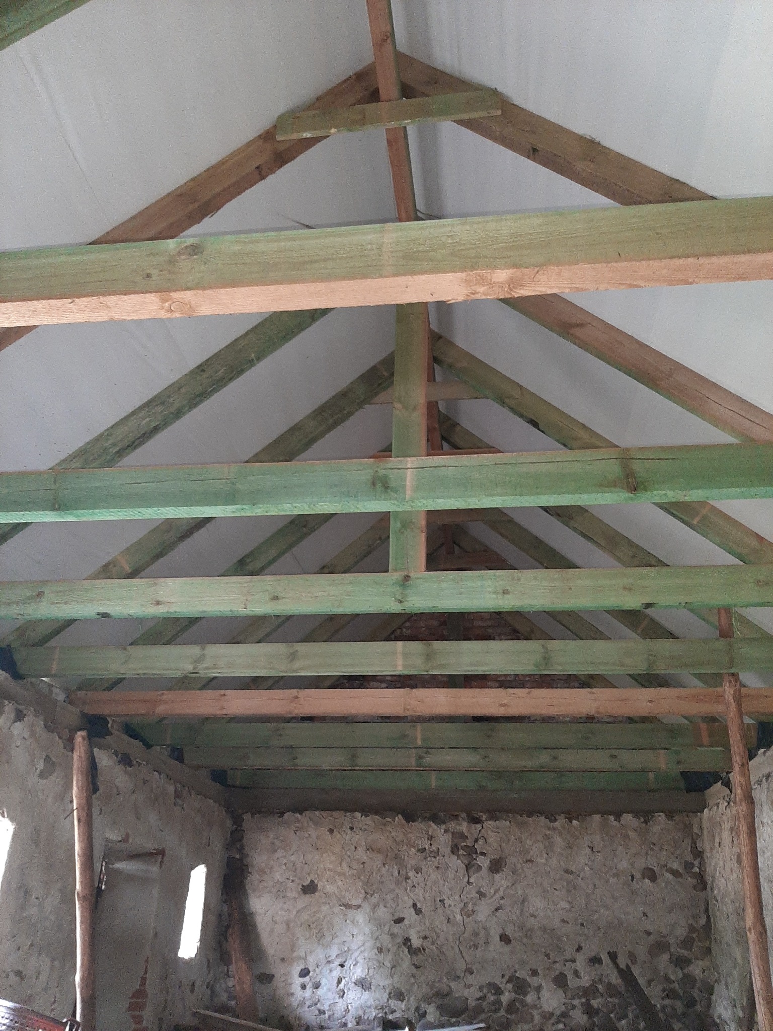 Wnętrze budynku w trakcie prac dekarskich, widoczne drewniane elementy konstrukcji dachu oraz surowa ściana z kamienia i cegły.