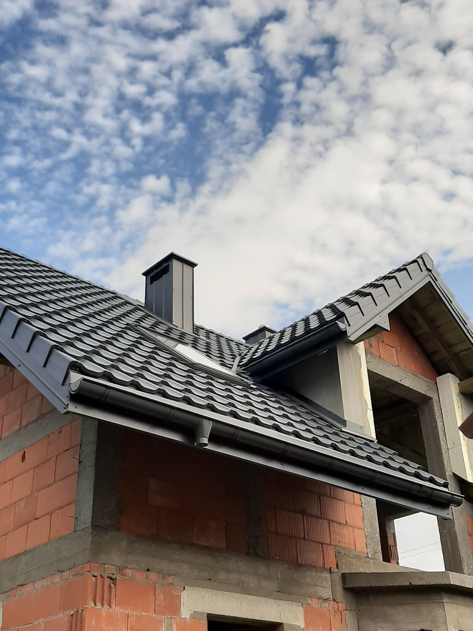 Fragment dachu z ciemnej blachodachówki z kominem na tle błękitnego nieba z obłokami, widoczna surowa konstrukcja budynku z czerwonej cegły.