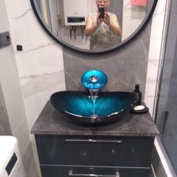 Uslugi remontowo-budowlane - Nowoczesna łazienka z umywalką w kolorze turkusowym, armaturą z wodospadem i odbiciem fotografa w lustrze.