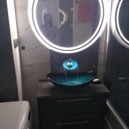 Uslugi remontowo-budowlane - Nowoczesna umywalka z podświetlaną wodą w łazience z okrągłym lustrem LED, widoczny fragment pralki z lewej strony.